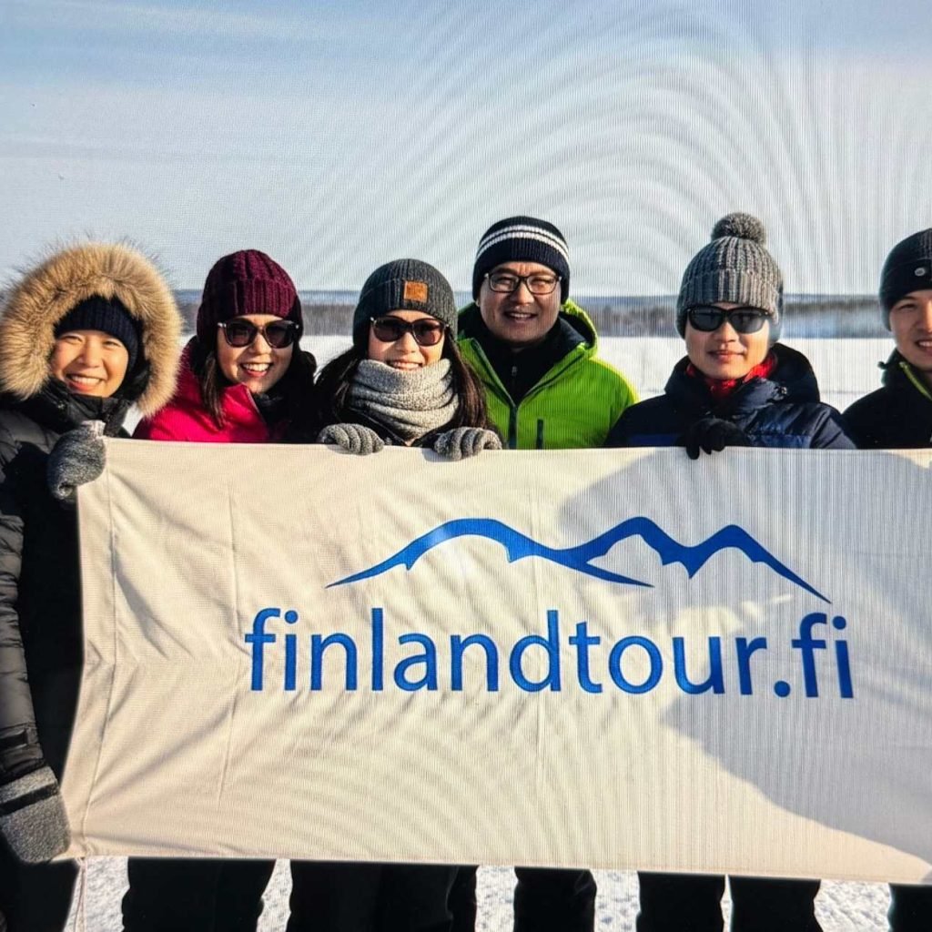 Group of Asian travelers at a snowmobile park in Finland holding a cloth banner with finlandtour.fi, during their 8 days Finland tour from Singapore