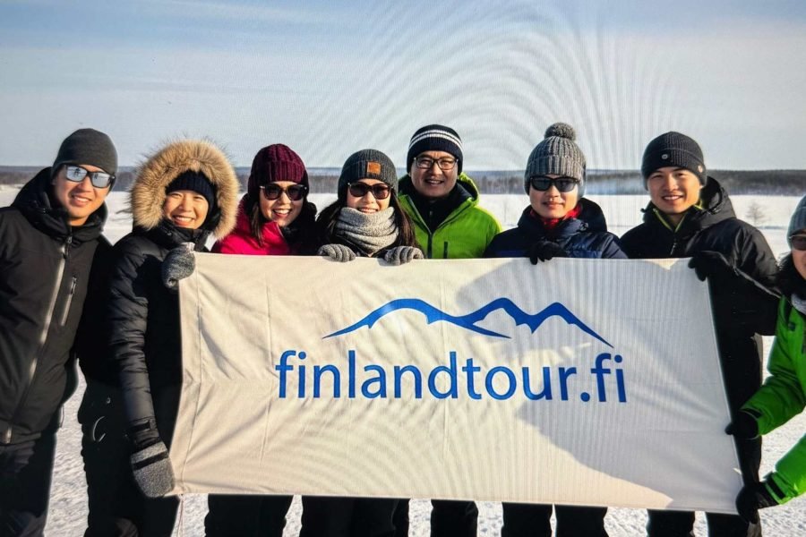 Group of Asian travelers at a snowmobile park in Finland holding a cloth banner with finlandtour.fi, during their 8 days Finland tour from Singapore
