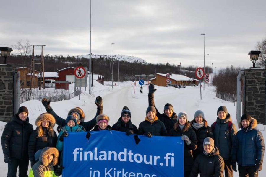 Group of happy travelers in Kirkenes holding a “finlandtour.fi in Kirkenes” banner during a 6 Day Rovaniemi, Saariselka & Kirkenes tour in Arctic Norway.