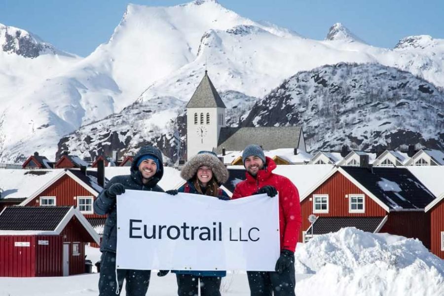 Travelers in Svolvær, Lofoten, holding an Eurotrail LLC banner with snow-covered mountains and waterfront cabins in the background during the 11D Helsinki, Saariselka, Rovaniemi, Kiruna, Narvik, Lofoten & Oslo tour.